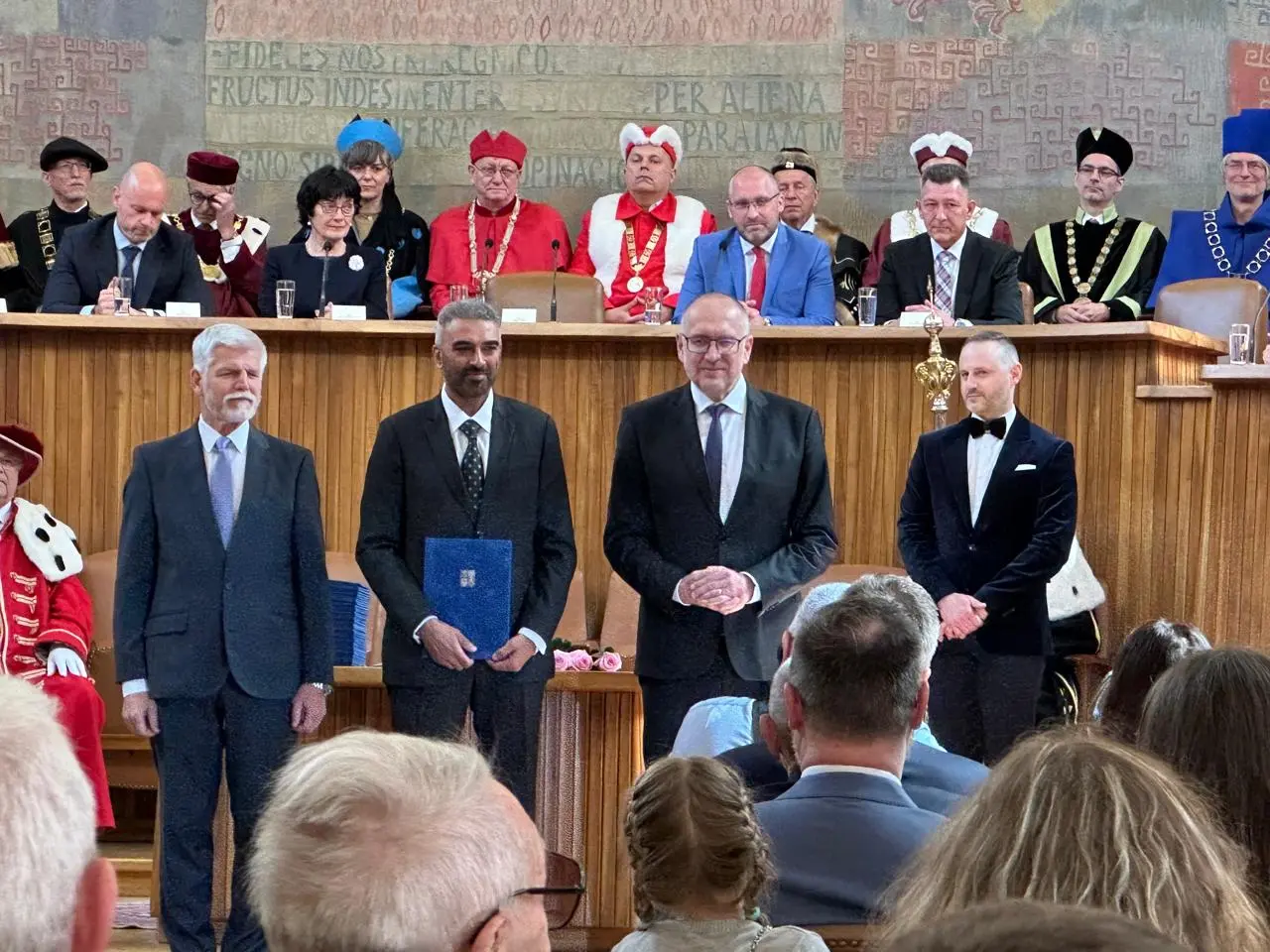 Four men standing in a university ceremonial hall, one holding a blue folder, with robed officials seated at a long table behind them.